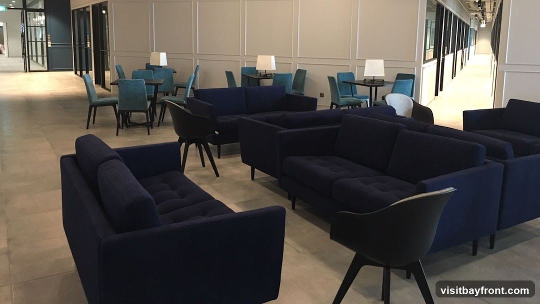 A contemporary lounge area with dark blue sofas and teal chairs arranged around small tables. The space features minimalist decor with white paneled walls and modern lamps on the tables. The flooring is a light-colored tile. In the background, glass-partitioned offices or meeting rooms are visible, suggesting a co-working space or office common area.
