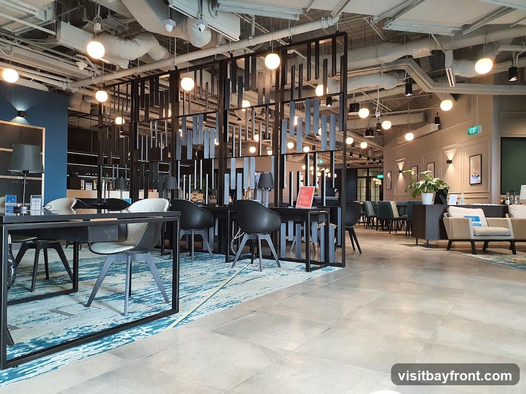 An interior view of a contemporary co-working space with a modern design. The space features a reception desk area with chairs and tables, separated by a decorative black partition. Further into the space, there are more seating areas with tables and chairs, and lounge seating. The ceiling is exposed with industrial-style lighting and ductwork. An 'EXIT' sign is visible on the wall.