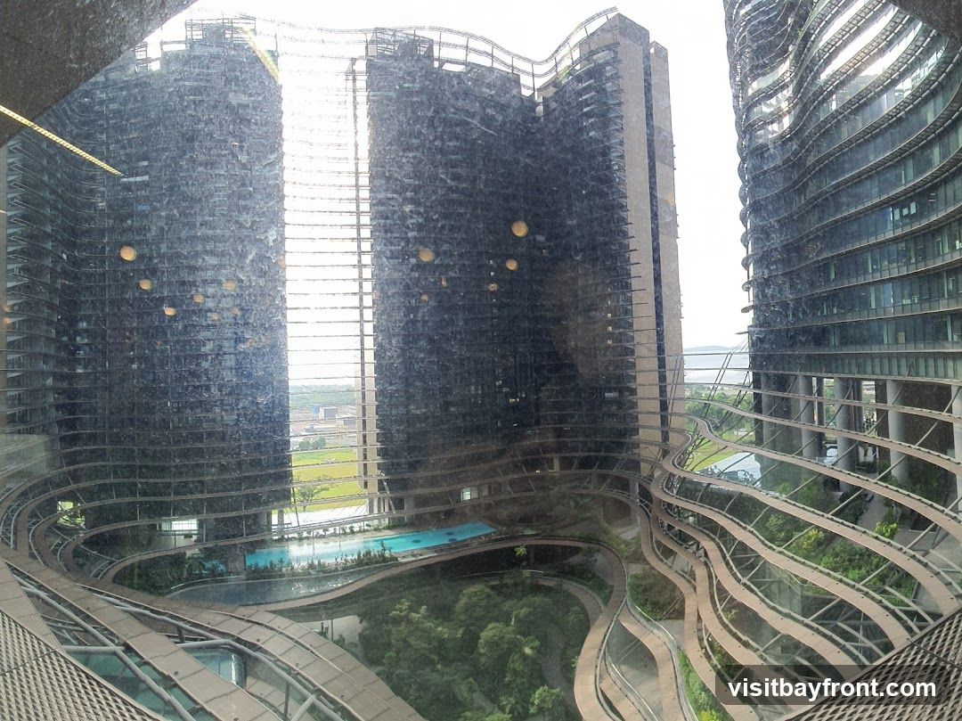 A view of several tall, modern buildings with distinctive wavy, textured facades. In the foreground, a curved walkway with a metallic railing winds through lush greenery. A swimming pool is visible in the mid-ground, surrounded by trees and landscaping. The sky is overcast.