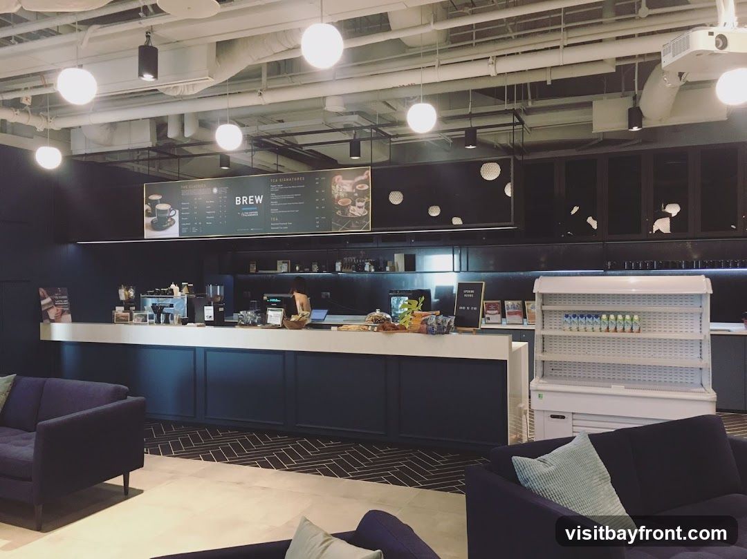 A view of a modern cafe interior with a long counter, seating areas, and a prominent menu board. The counter displays coffee machines, pastries, and drinks. Two dark blue sofas are visible in the foreground. The menu board above the counter features 'BREW' and 'TEA SIGNATURES' with various drink options and prices.
