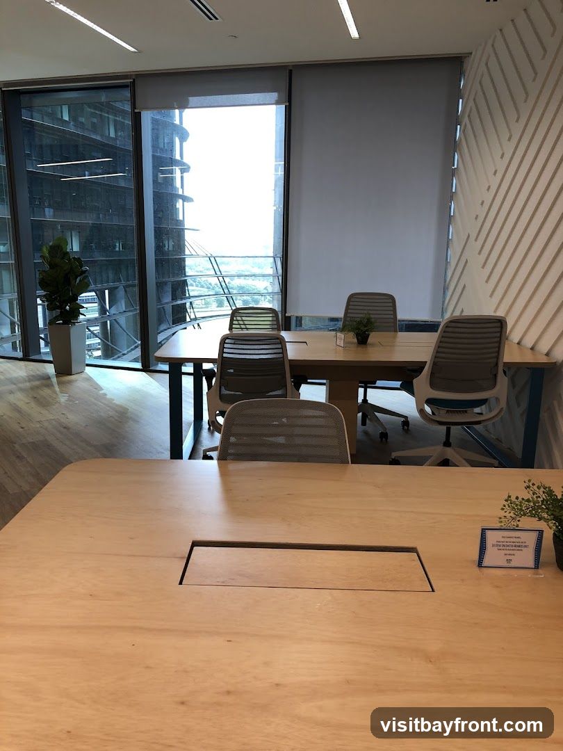 A contemporary meeting room with large windows offering city views. The room features several wooden tables and chairs, a potted plant, and blinds covering some of the windows. A small sign is visible on one of the tables.