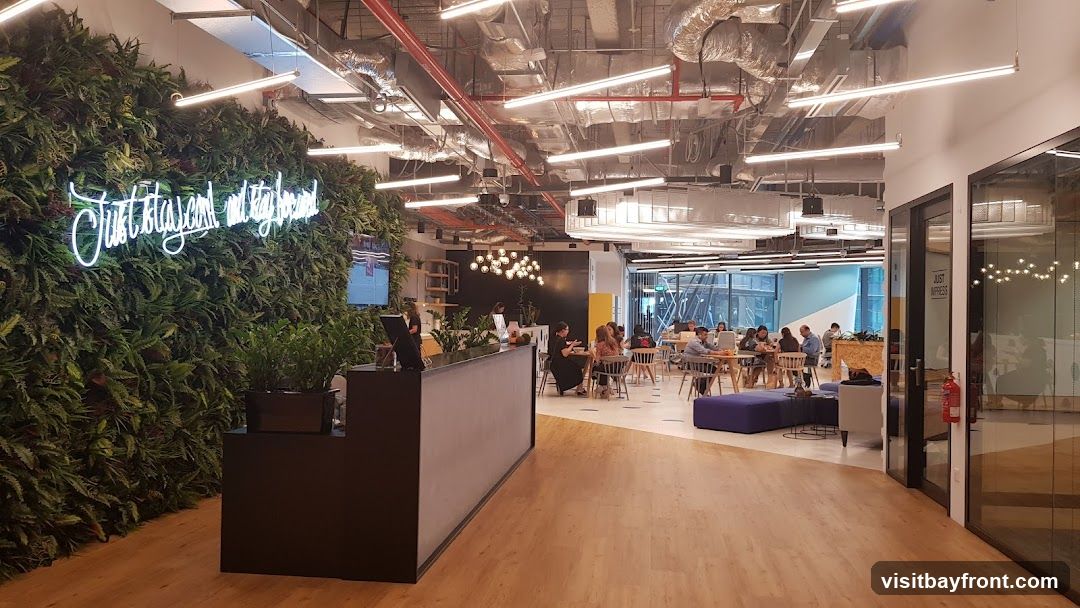 An interior view of a modern office space featuring a prominent living green wall adorned with a white neon sign that reads "Just stay cool and stay inspired." A reception desk is situated in the foreground. The background reveals a co-working area with people seated at tables, engaged in work and conversation. The ceiling displays exposed ductwork and industrial lighting, contributing to the contemporary aesthetic.