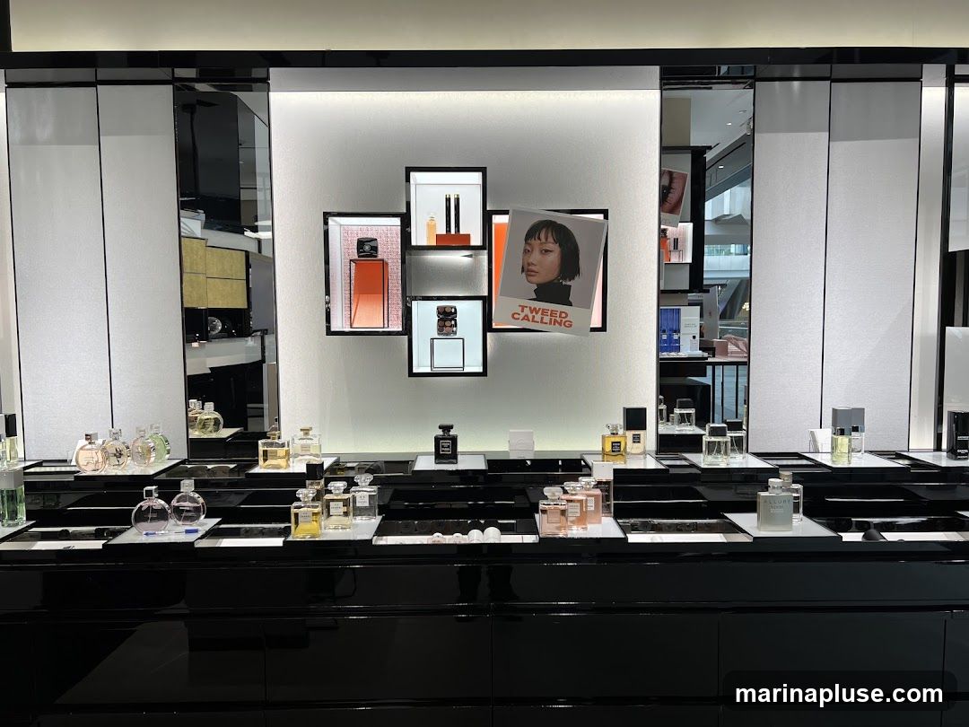 A well-lit display of various perfumes and cosmetics on a black, multi-tiered counter. Behind the counter, a wall features framed product images and a poster with the text "TWEED CALLING" and a portrait of a woman. The display includes numerous perfume bottles of different shapes and sizes, as well as some makeup products.