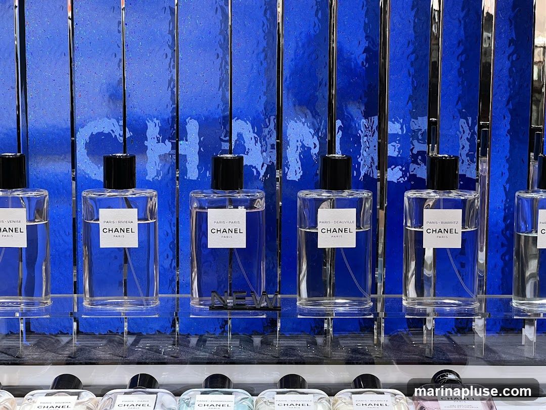 A display of Chanel perfumes with blue, textured background. The bottles are clear glass with black caps and white labels. The labels indicate different scents such as Paris-Venise, Paris-Riviera, Paris-Deauville, and Paris-Biarritz. The Chanel logo is visible on the background.