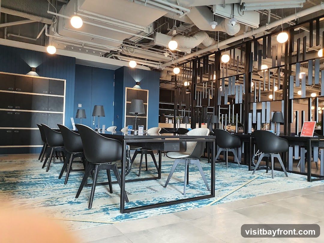 A contemporary co-working space with dark blue walls, black furniture, and a blue and white patterned rug. The space features multiple desks with chairs, each equipped with a desk lamp. Overhead, exposed pipes and modern light fixtures are visible. The overall ambiance is sleek and professional.