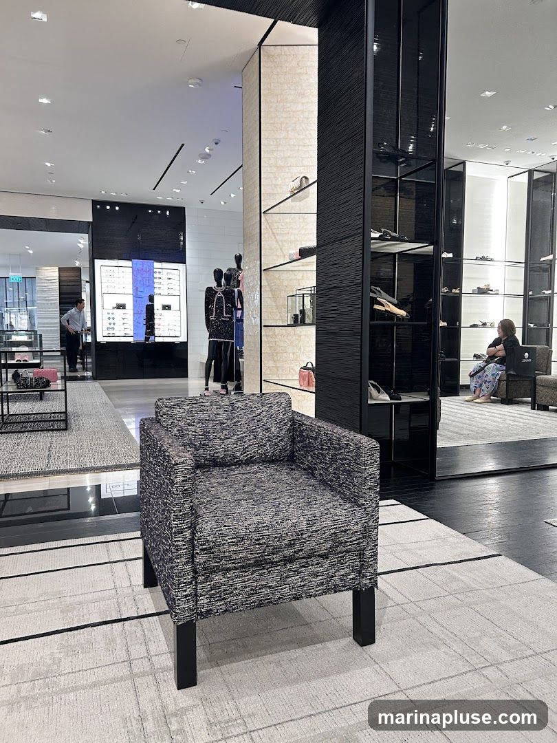 A plush armchair sits on a patterned rug in a high-end retail store. In the background, mannequins display designer clothing, and shelves showcase handbags and shoes. A customer is seated in the distance. The store features sleek displays and modern lighting.