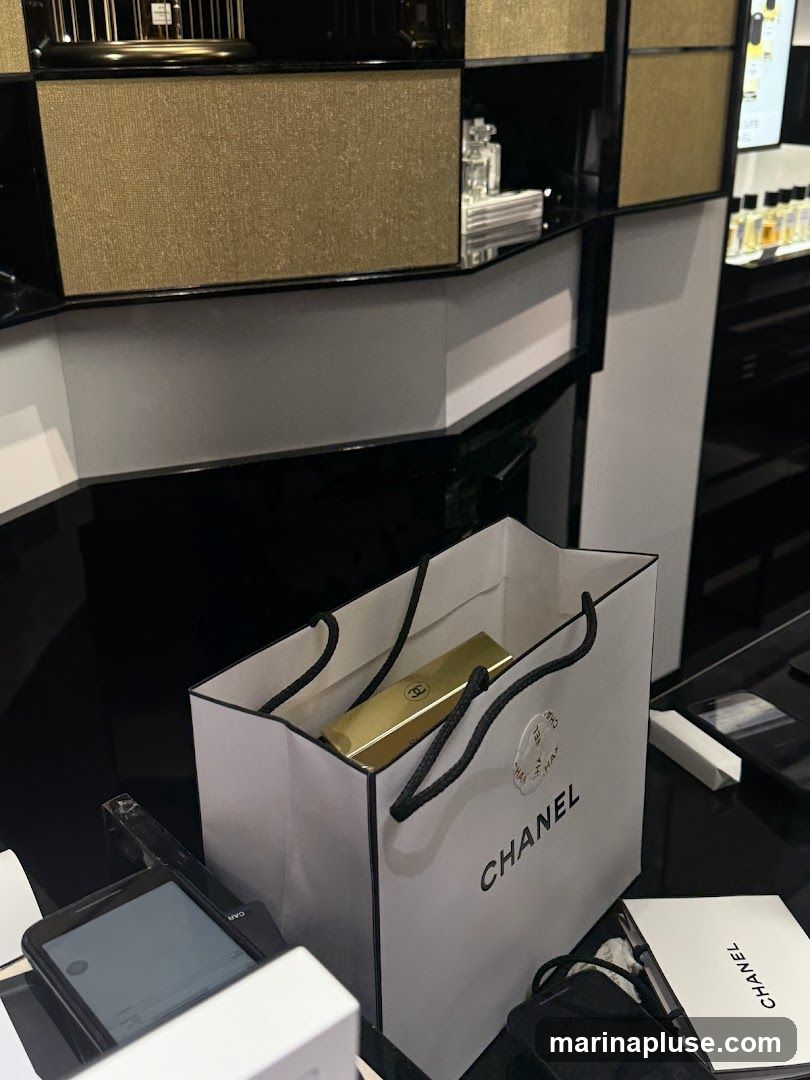 A white Chanel shopping bag with black handles contains a gold lipstick. The bag has the Chanel logo printed on it. In front of the bag is a smartphone and other items. In the background, there are shelves displaying perfume bottles and other products.