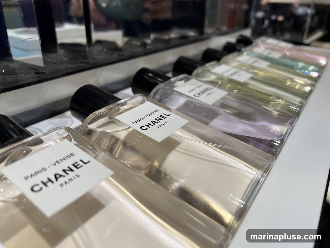 A close-up, angled shot showcases a row of Chanel perfume bottles displayed on a white surface. The bottles are clear glass with black caps and white labels. The labels indicate different scents, with "PARIS - VENISE" and "PARIS - RIVIERA" being visible. The perfumes inside vary in color, from clear to pale yellow, lavender, and pink. The background is blurred, suggesting an indoor retail environment.