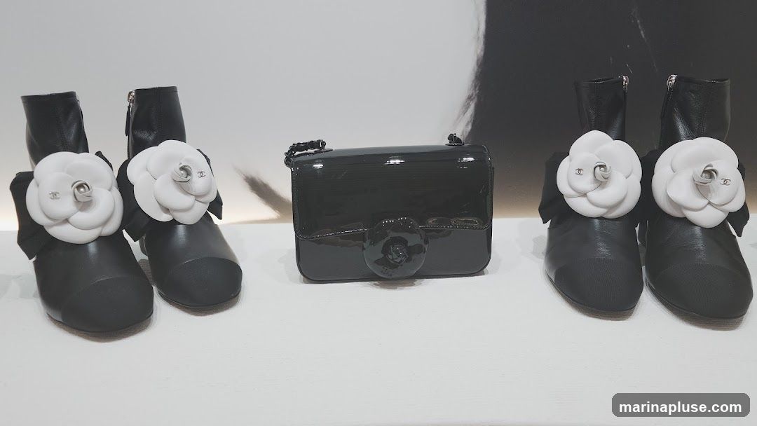 A black patent leather handbag with a camellia embellishment is displayed between two pairs of black ankle boots. Each boot is adorned with a large white camellia flower with black ribbon accents and a small Chanel logo.