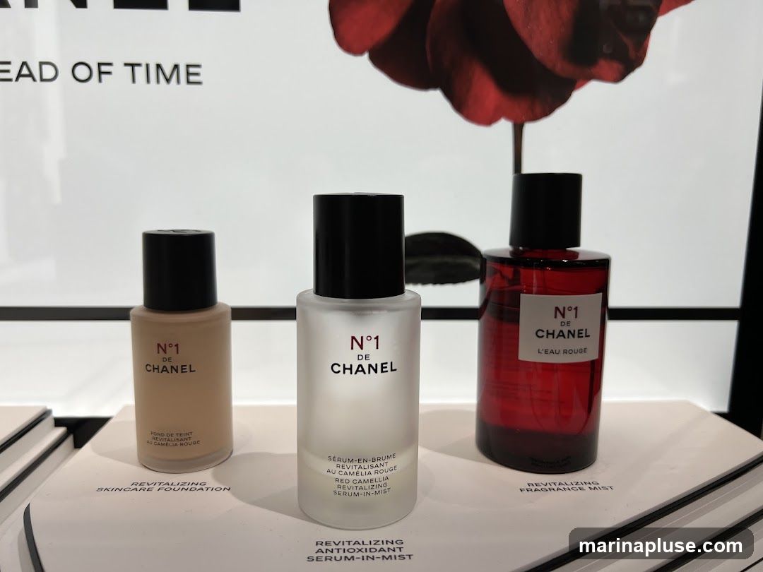 Three bottles of Chanel N°1 de Chanel products are displayed on a white surface. The products include a revitalizing skincare foundation, a revitalizing antioxidant serum-in-mist, and a revitalizing fragrance mist. The background features a large image of a red rose and text that reads "EAD OF TIME".