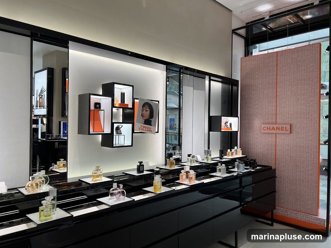 A well-lit interior display of Chanel perfumes and beauty products. Various bottles of perfume are arranged on sleek black shelves. A large textured wall panel with the Chanel logo is visible on the right. A display shelf features framed images and product samples.