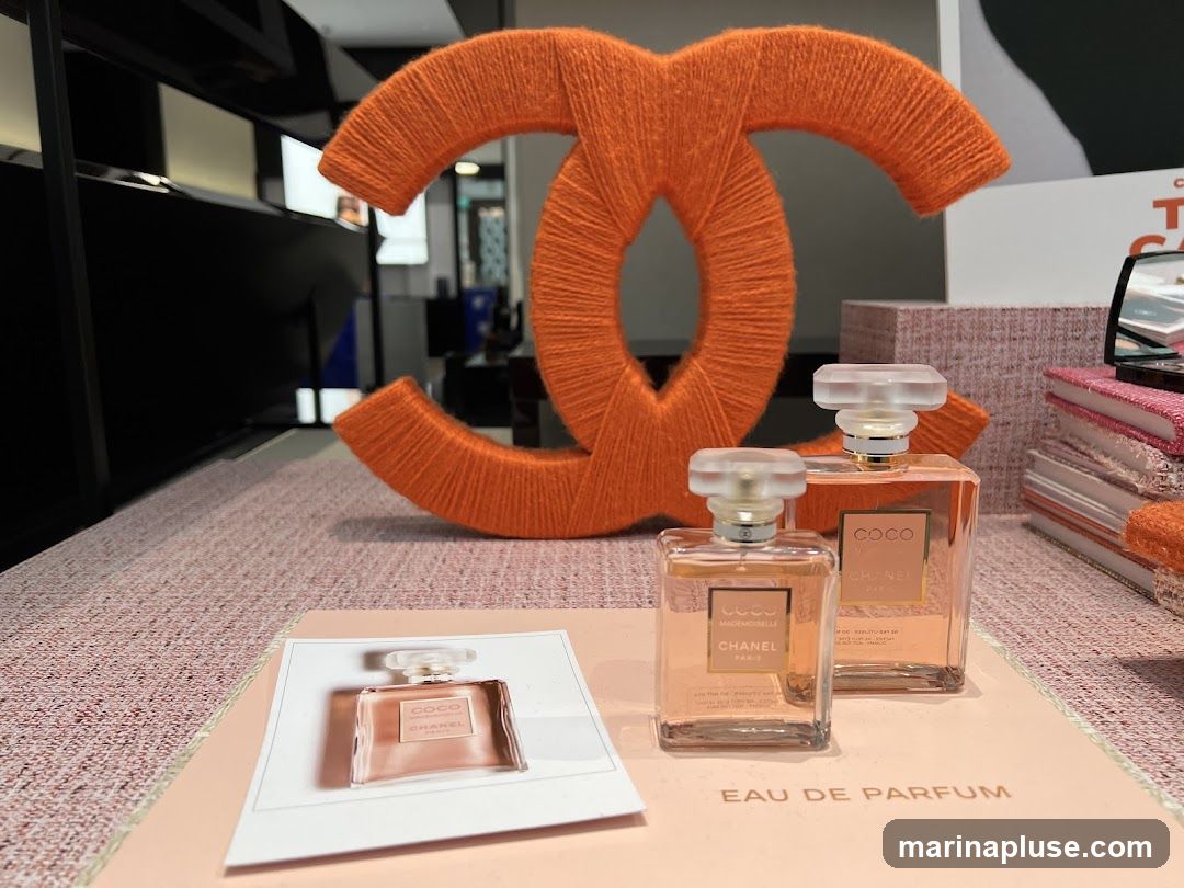 A display featuring two bottles of Chanel Coco Mademoiselle Eau de Parfum and a sample card. In the background, a large orange Chanel logo made of yarn is visible, along with stacked pink and white boxes and a compact mirror. The display appears to be set up on a textured pink surface.