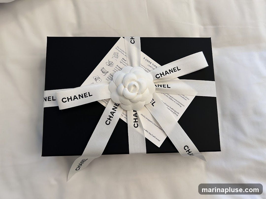 A black Chanel gift box is tied with a white ribbon featuring the Chanel logo. A white camellia flower adorns the ribbon. Care instructions are visible beneath the ribbon.