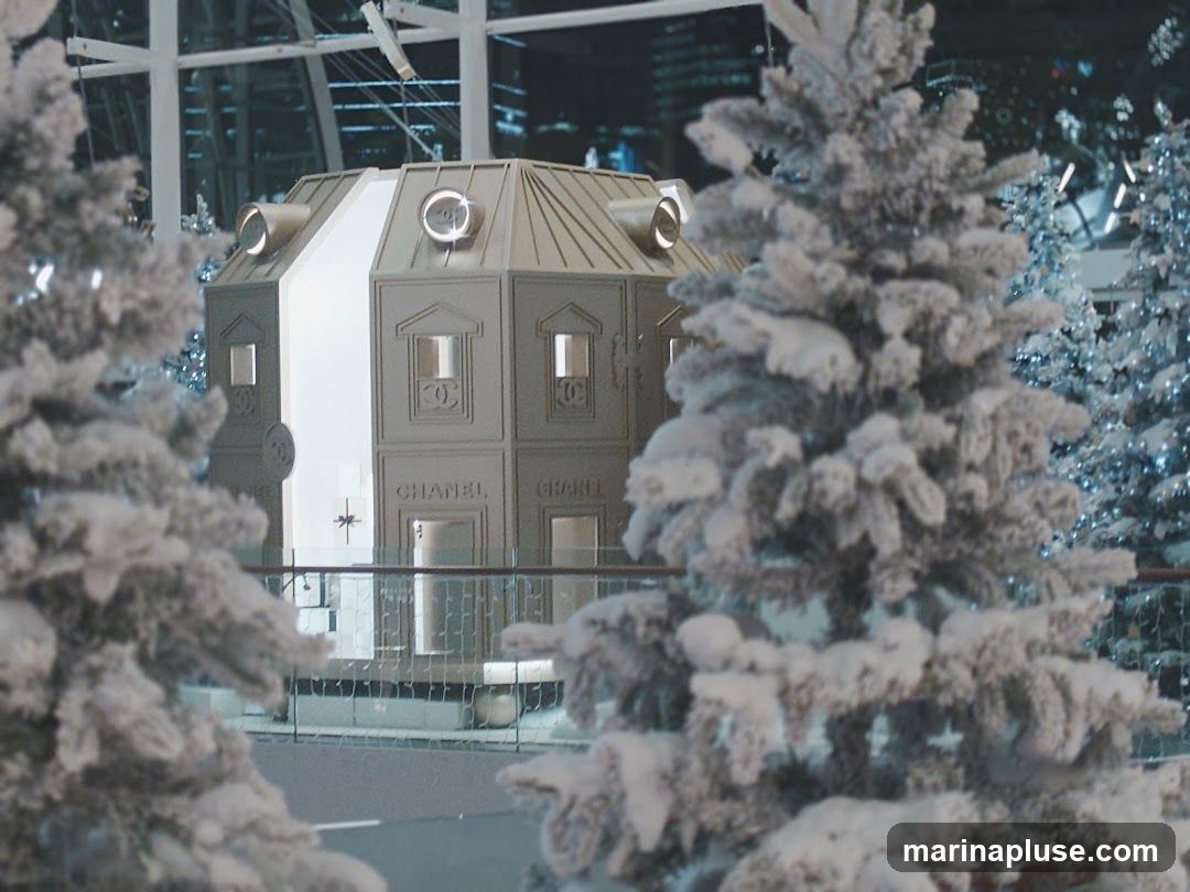A miniature Chanel-themed house is displayed amongst snow-covered Christmas trees. The house features the Chanel logo and name on its facade, resembling a storefront. The background appears to be an indoor setting with architectural elements and lights.
