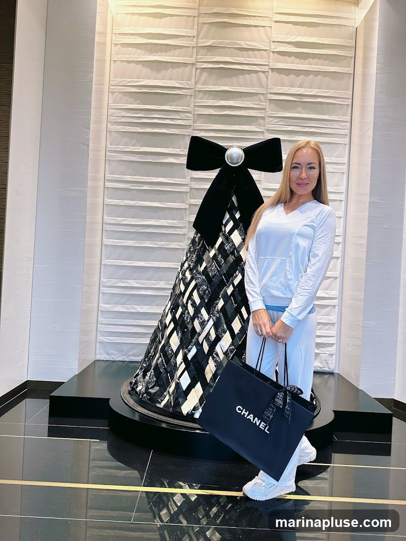 A woman stands in front of a display featuring a large black bow and a black and white patterned skirt-like structure. She is holding a black Chanel shopping bag. The background has a textured white wall.
