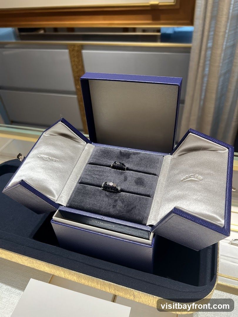 A blue velvet-lined jewelry box is open, displaying two rings. The box has satin lining inside the lid, with embossed logos. The rings appear to be made of dark metal and have intricate designs. The box is placed on a textured gold tray, with a blurred background suggesting a retail environment.
