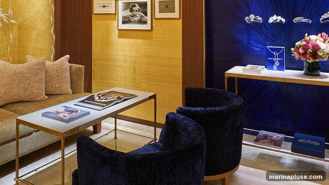 A plush seating area with a sofa and two armchairs is arranged around a coffee table. On the table are two magazines or books. A display shelf on the right showcases several tiaras, a bouquet of flowers, and branded items including a blue box with the word 'CHAUMET' and a necklace illustration. Below, another shelf displays more books and a jewelry box. The walls are decorated with textured gold wallpaper and framed artwork, contrasting with a vibrant blue backdrop behind the display shelf.