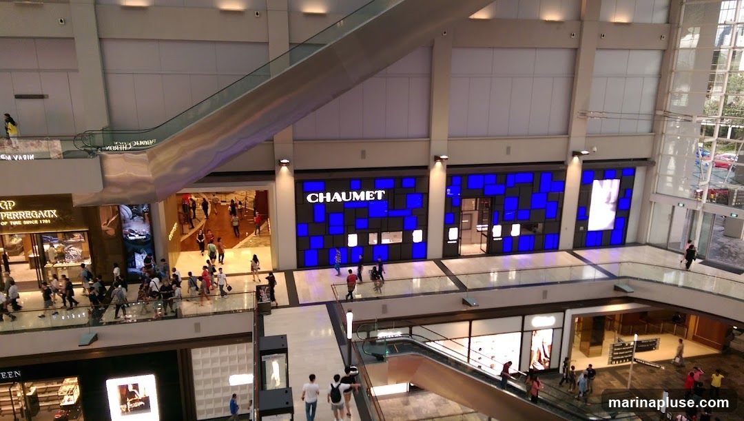 The image shows the interior of a multi-level shopping mall. In the center, a store with the name "CHAUMET" is visible, featuring a facade with blue and white square panels. To the left, another store with the name "GP PERREGAUX" is displayed, showcasing watches. Escalators and walkways are present, with shoppers moving around. The overall ambiance is bright and modern, typical of a high-end retail environment.