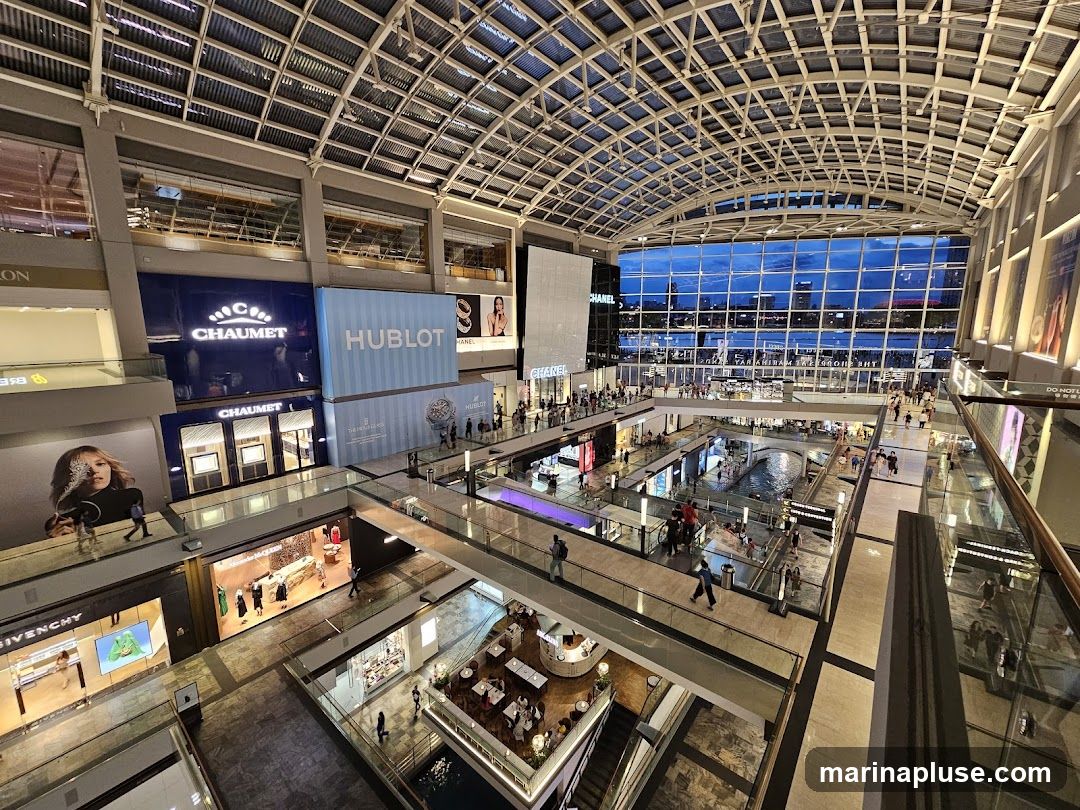 A wide-angle view of the interior of a luxurious shopping mall. The mall features multiple levels with walkways and escalators, overlooking a central canal with boats. Large storefronts with prominent brand names like CHAUMET, HUBLOT, GIVENCHY, and CHANEL are visible. The ceiling is a complex glass and metal structure, and the far end of the mall has a large glass facade offering a view of the city skyline at dusk.