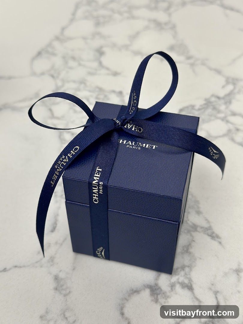 A dark blue gift box from Chaumet Paris, tied with a navy blue ribbon. The ribbon has the Chaumet Paris logo printed on it in gold lettering. The box is placed on a marble surface.