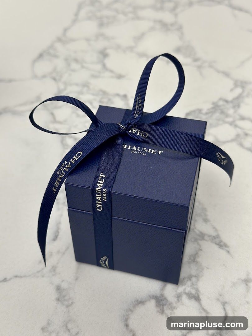 A dark blue gift box from Chaumet Paris, tied with a navy blue ribbon. The ribbon has the Chaumet Paris logo printed on it in gold lettering. The box is placed on a marble surface.