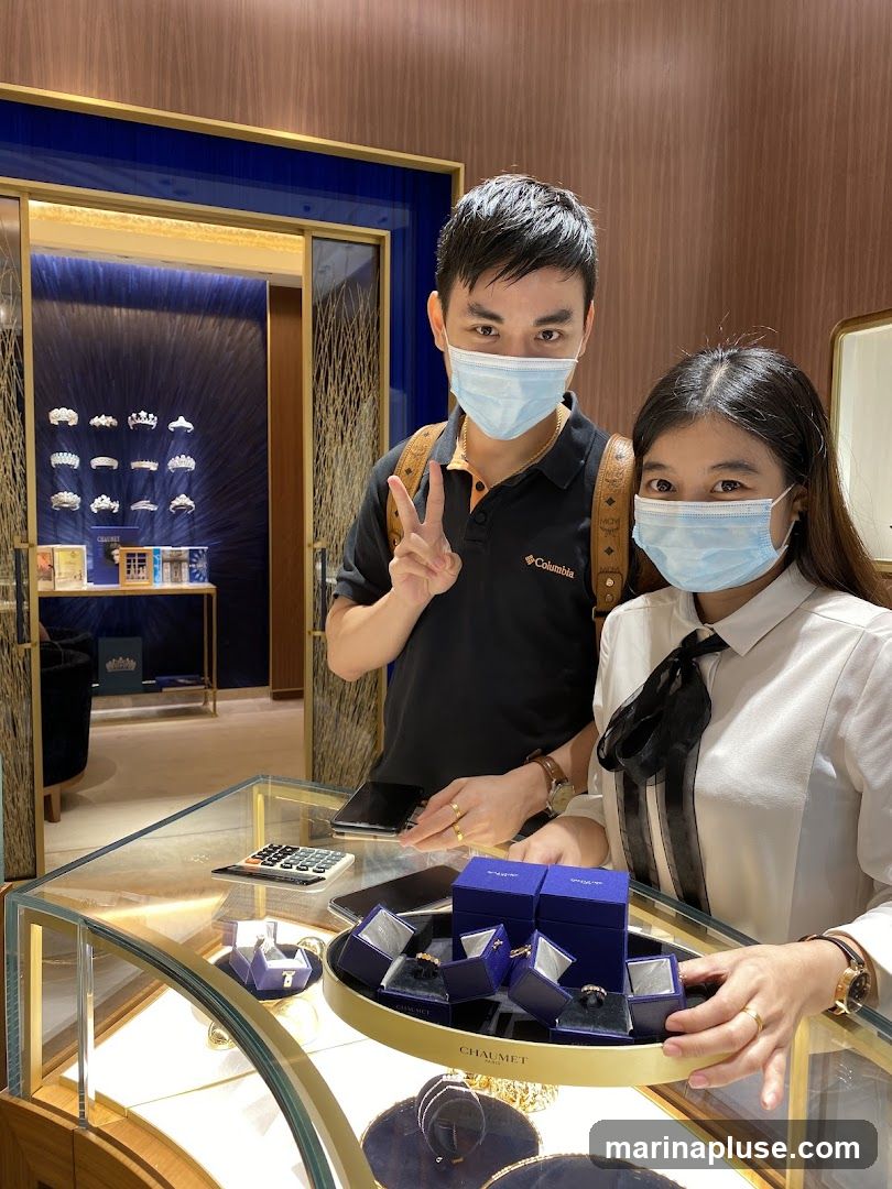 A man and a woman wearing face masks are standing in a jewelry store. The man is wearing a black polo shirt and the woman is wearing a white collared shirt. They are standing behind a glass display case filled with jewelry boxes and rings. In the background, there are displays of tiaras and other jewelry. The store appears to be upscale.