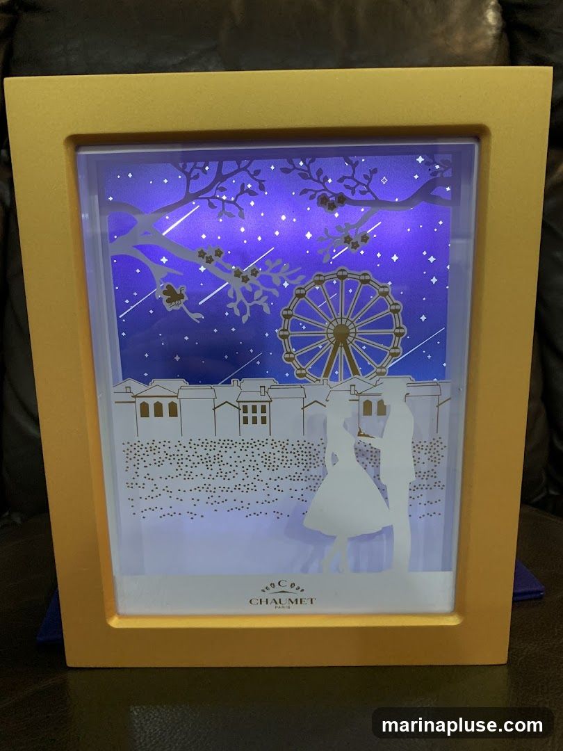 A golden-framed light-up diorama depicts a romantic scene under a starry night sky. Silhouettes of a couple embracing are in the foreground, with a Ferris wheel and a cityscape in the background. The diorama is illuminated from behind, casting a soft glow on the intricate cutouts. The brand name "CHAUMET PARIS" is visible at the bottom center.