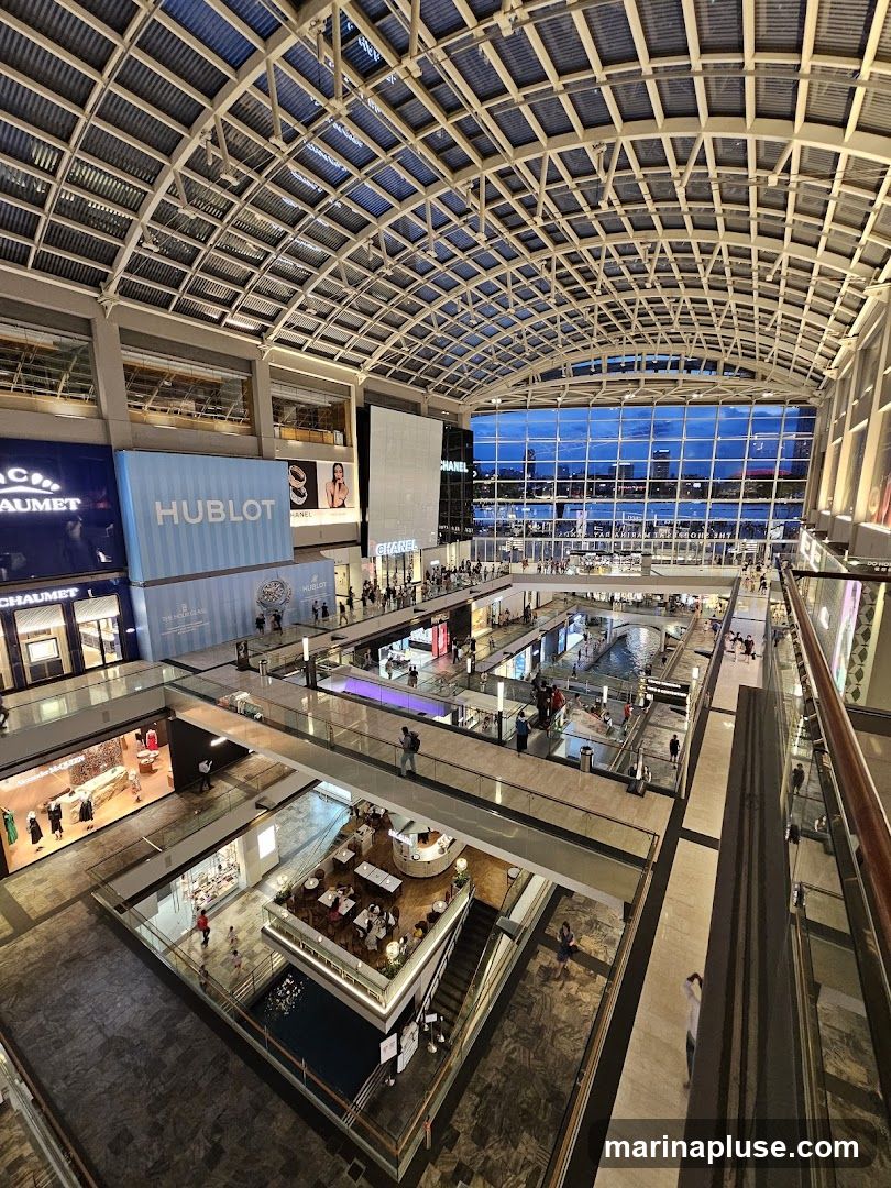 An aerial view of the interior of a multi-level shopping mall with a large glass roof. Shops such as Chanel and Hublot are visible. There is a central canal or water feature and several escalators and walkways connecting the different levels. People are seen browsing and walking around the mall.