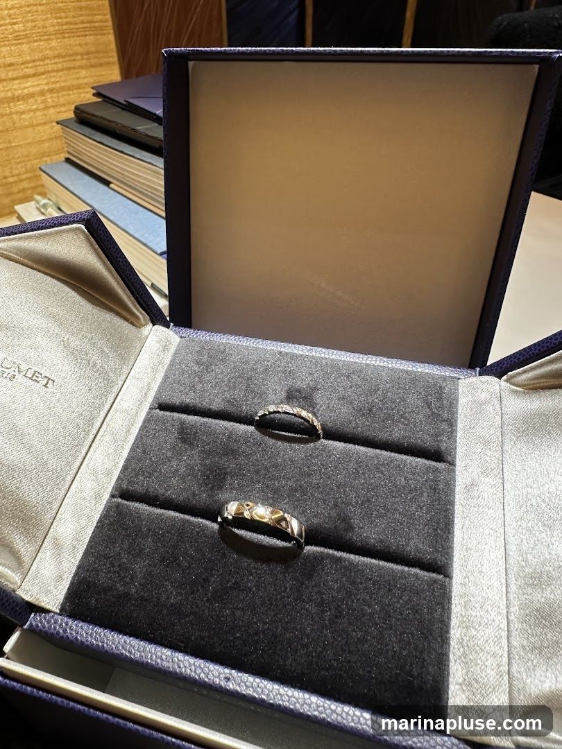 A dark blue velvet-lined jewelry box is open, displaying two rings. The top ring is delicate and appears to be paved with small stones. The bottom ring is wider and has a geometric, interlocking design. The inside lid of the box has a satin finish and a faint logo that reads "CHAUMET PARIS". A stack of books is visible in the background.