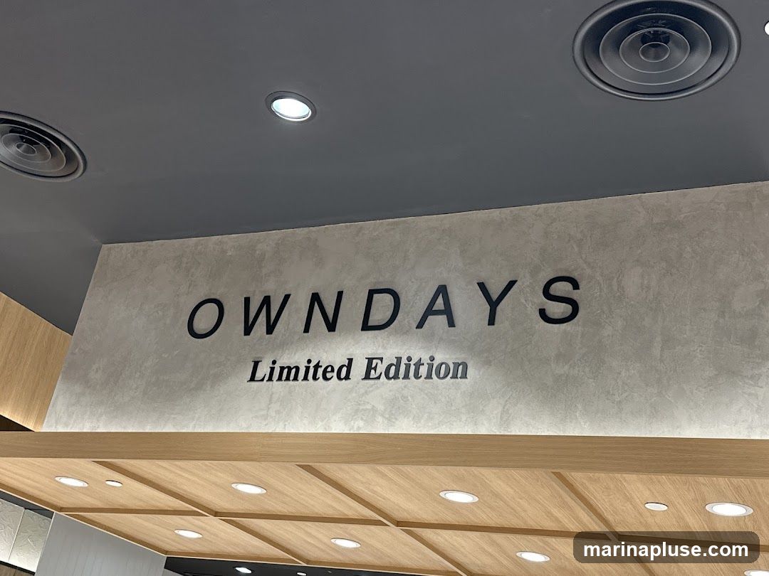 A close-up shot of a sign that reads "OWNDAYS Limited Edition" mounted on a textured wall. The sign is in black lettering, with "OWNDAYS" in a larger, bold font and "Limited Edition" in a smaller, italicized font below it. The ceiling above is dark gray with recessed lighting and air vents. Below the sign, a wooden paneling with square insets and more recessed lighting is visible.