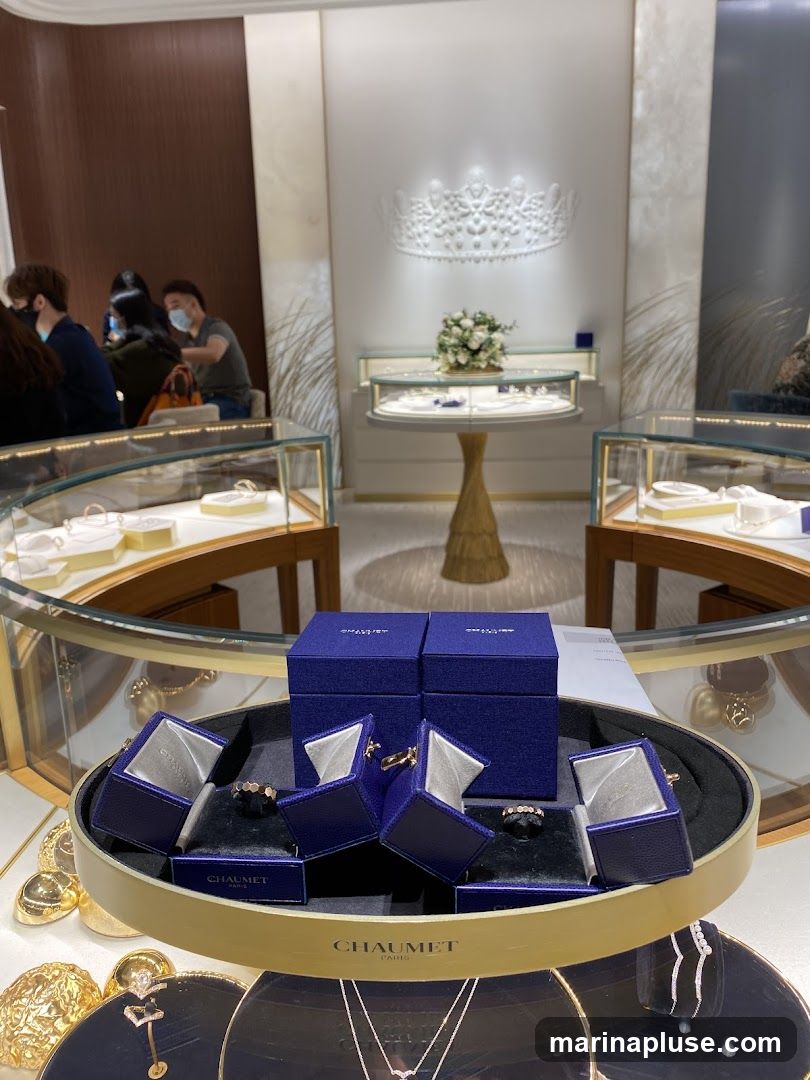 A display of luxury jewelry in a store. In the foreground, several open blue jewelry boxes are arranged on a circular tray, with rings visible inside some of them. The tray has the CHAUMET PARIS logo. Behind this, more display cases with jewelry are visible. In the background, a large white crown-like sculpture adorns the wall above a display table with a floral arrangement. Several customers are seated in the background, appearing to be served by staff.
