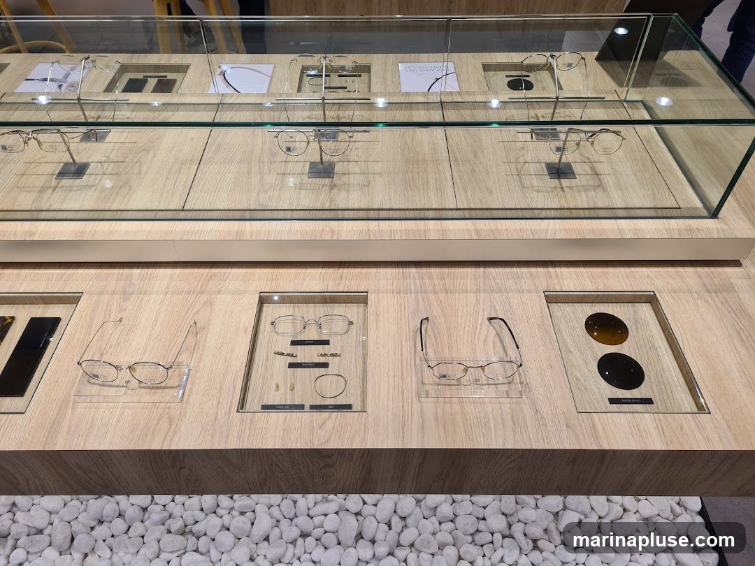 A display case filled with various eyeglasses and sunglasses. The display is made of wood with glass shelves and compartments. Some of the eyewear is presented on stands, while others are laid flat. There are also samples of lenses shown. The background is blurred, but appears to be an indoor retail environment.