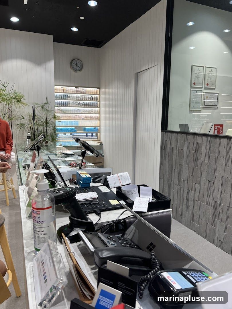 A counter area with various items, including a hand sanitizer bottle, a barcode scanner, a laptop, a telephone, payment terminals, and stacks of receipts. Behind the counter, shelves are stocked with numerous boxes, possibly cigarettes or similar products. Several framed certificates are displayed on the wall in the background.