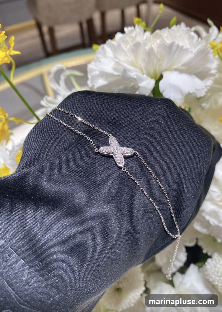 A close-up shot of a delicate silver bracelet with a diamond-encrusted clover-shaped charm. The bracelet is held by a gloved hand, with a blurred background of white and yellow flowers.