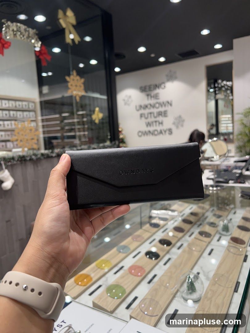 A hand holds a black glasses case with the logo "OWNDAYS" embossed on it. In the background, a display case shows various colored lenses and clear lenses, arranged on wooden stands. The store appears to be decorated for the holidays with snowflakes and bows. A wall behind the display has text that reads "SEEING THE UNKNOWN FUTURE WITH OWNDAYS.".