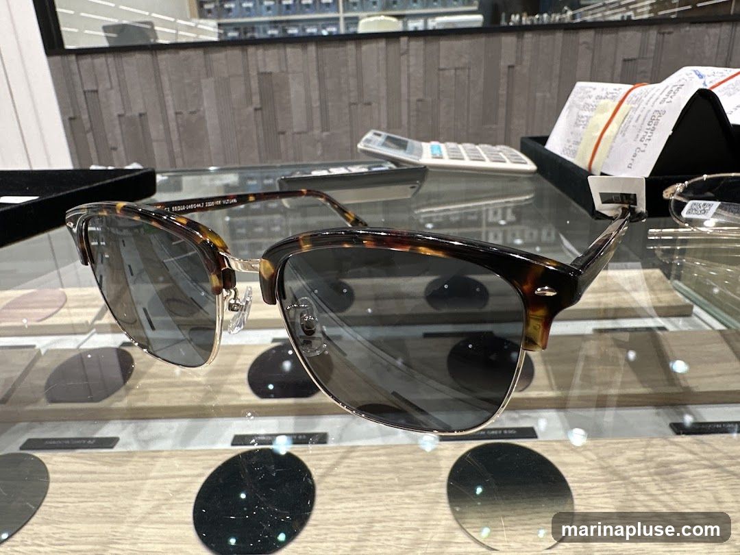 A pair of tortoiseshell sunglasses with gold accents is displayed on a glass surface. In the background, a calculator and receipts are visible, along with shelves of eyewear and lens samples.