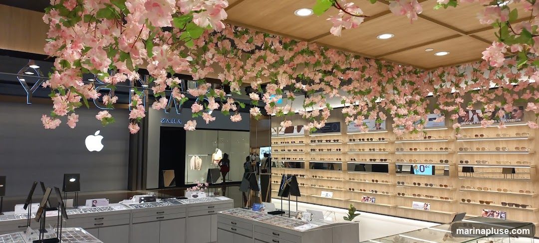 The interior of an eyewear store is decorated with artificial cherry blossoms hanging from the ceiling. Shelves are filled with various pairs of glasses and sunglasses. In the background, there is a Zara store and an Apple logo.