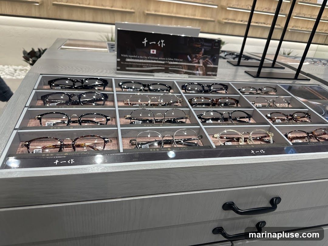 A display case filled with various styles of eyeglasses. The glasses are arranged in rows and compartments on a light gray wooden surface. Some glasses have tortoiseshell frames, while others have metal frames. A sign with Japanese text and an image of a person wearing glasses is visible in the background.