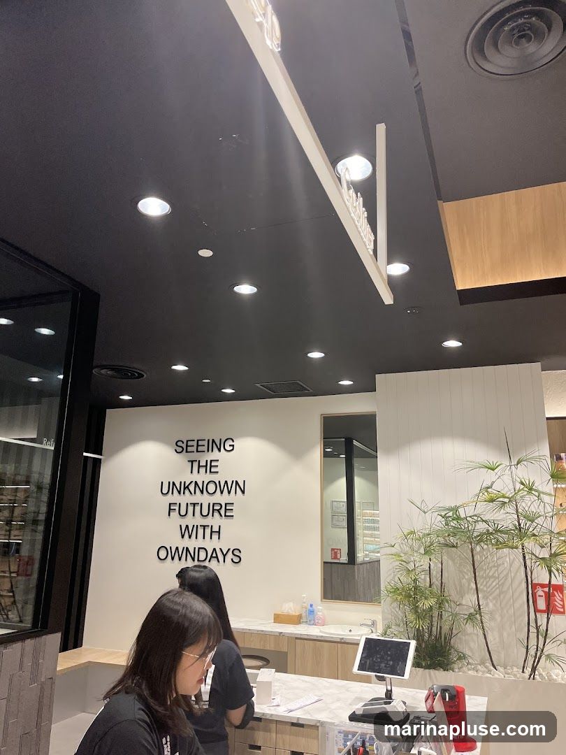The image shows the interior of a store. A large quote on the wall reads "SEEING THE UNKNOWN FUTURE WITH OWNDAYS". Two women are visible behind a counter. There is a tablet on the counter, along with a red card reader. Plants are placed to the right of the counter. A sign with the store's name is visible hanging from the ceiling.