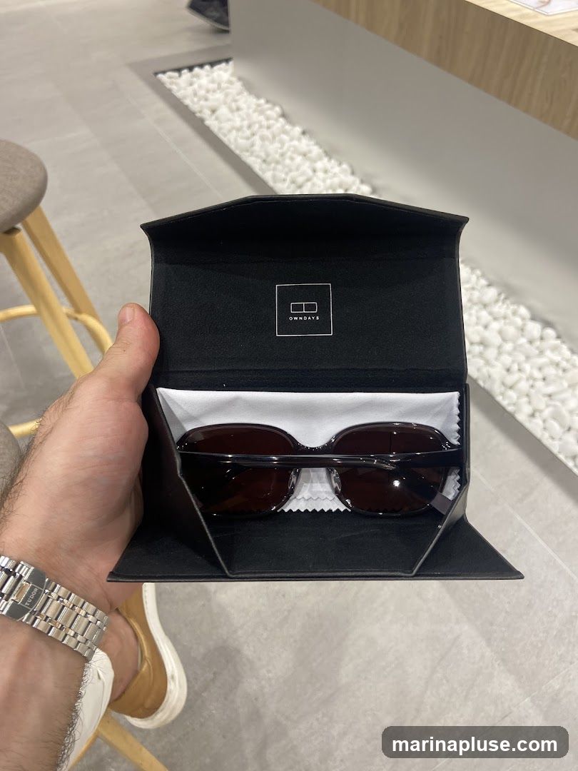 A person's hand holds open a black case containing a pair of dark-rimmed sunglasses. The case has a white logo with the text "OWNDAYS" inside a square. The background shows a modern interior with grey flooring and white decorative stones.