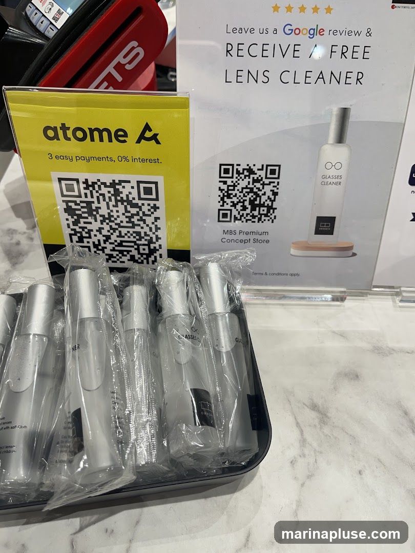 A display featuring a yellow sign with the Atome logo and QR code for 3 easy payments, 0% interest. Next to it, a white sign advertises a free lens cleaner with a Google review, showing a bottle of glasses cleaner and a QR code for MBS Premium Concept Store. A tray of individually wrapped lens cleaner bottles is in the foreground.