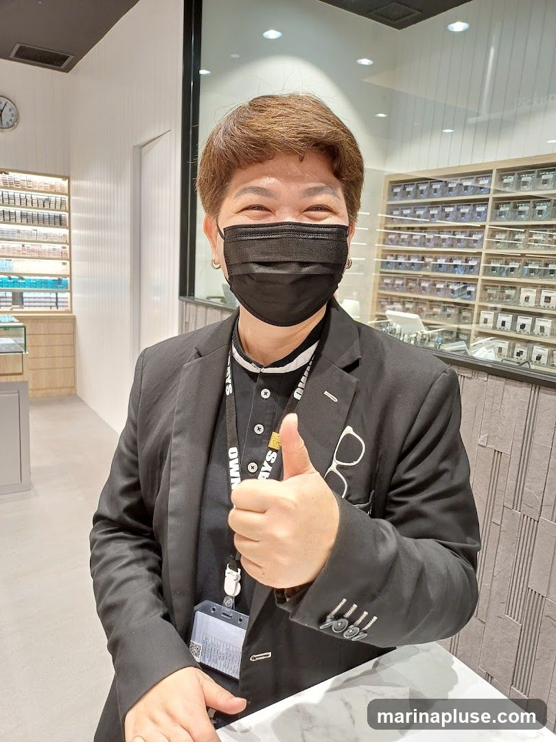 A person wearing a black suit and a black face mask gives a thumbs up while smiling. They are wearing a lanyard with an ID badge. In the background, there are shelves filled with what appear to be contact lenses or small optical items.