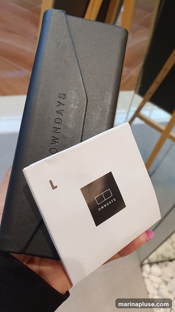 A hand holds a dark gray, textured eyewear case with the embossed word "OWNDAYS" visible. In front of the case is a white booklet with a black square logo containing the word "OWNDAYS" and two adjacent rectangles. The background appears to be an interior space with wooden beams and reflective surfaces.