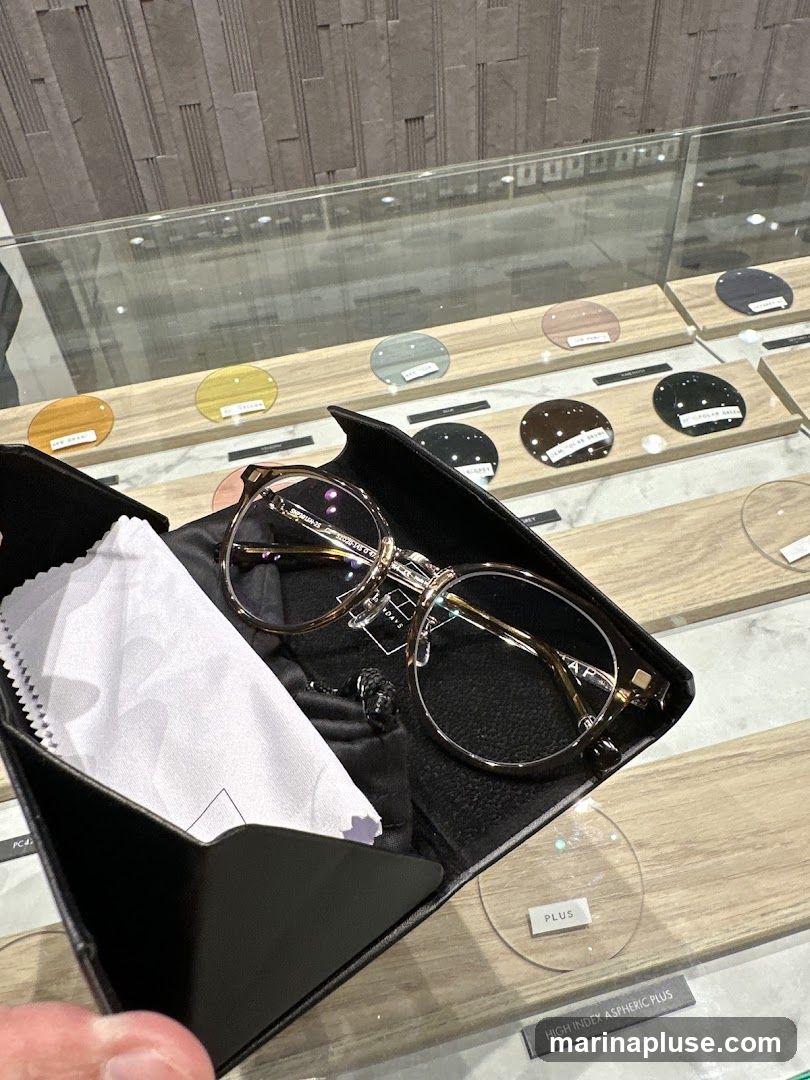 A pair of eyeglasses in a black case with a cleaning cloth. Behind the case, various lens samples are displayed on wooden stands, with labels indicating different types and colors.