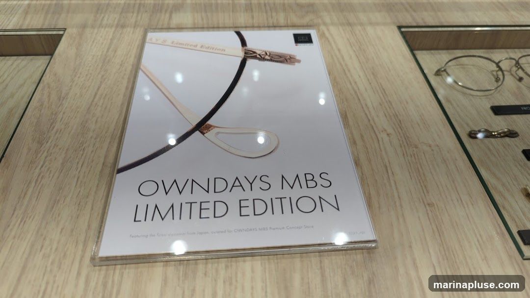 A display featuring a sign for OWNDAYS MBS Limited Edition eyewear. The sign shows a close-up of a pair of glasses and text. To the right, a display case shows several pairs of glasses.