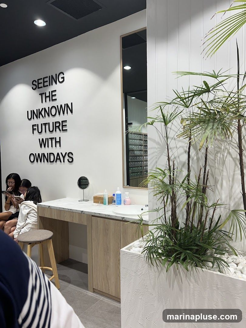 The image shows the interior of a modern retail space, possibly an optician or eyewear store. A large mirror reflects a wall lined with shelves displaying numerous items, likely glasses or contact lenses. On the left, a white wall features large black lettering that reads "SEEING THE UNKNOWN FUTURE WITH OWNDAYS." Below this text is a vanity area with a marble countertop, a sink, and various bottles and a small mirror. Three young women are seated at the vanity, looking at their phones. To the right, a large white planter contains lush green plants, adding a natural element to the clean, minimalist design.