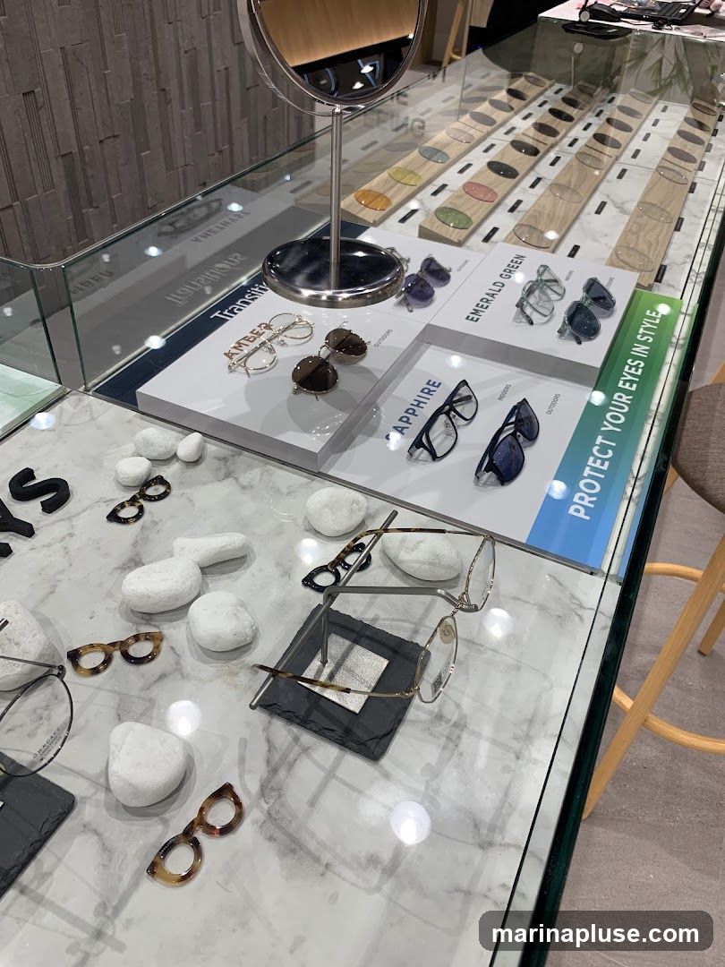 A display case filled with various eyeglasses and lenses. Different styles of frames are showcased on white stands, with labels such as "AMBER", "EMERALD GREEN", and "SAPPHIRE". A selection of colored lenses is arranged in the background. The display also features decorative white stones and a mirror. A sign reads "PROTECT YOUR EYES IN STYLE".