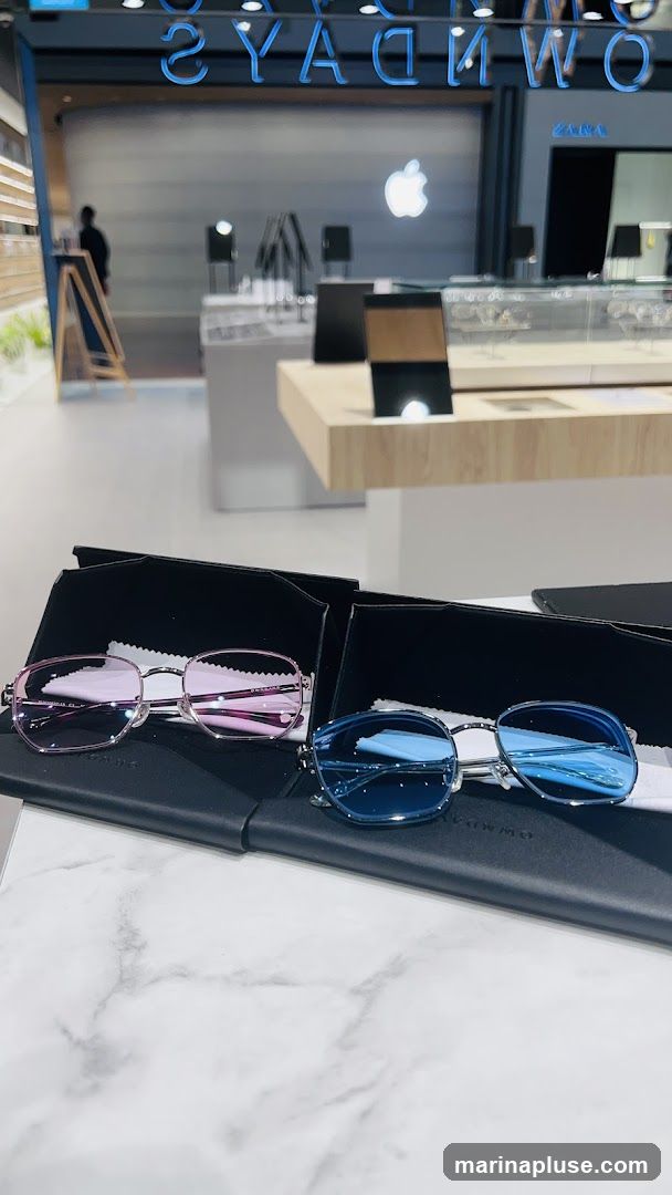 Two pairs of eyeglasses, one pink and one blue, are displayed on black cases on a marble surface. In the background, an Apple logo is visible, along with neon blue lettering that appears to spell out "SUNDAYS" and "ZARA". The setting suggests an optical shop or a retail store with an electronics section.