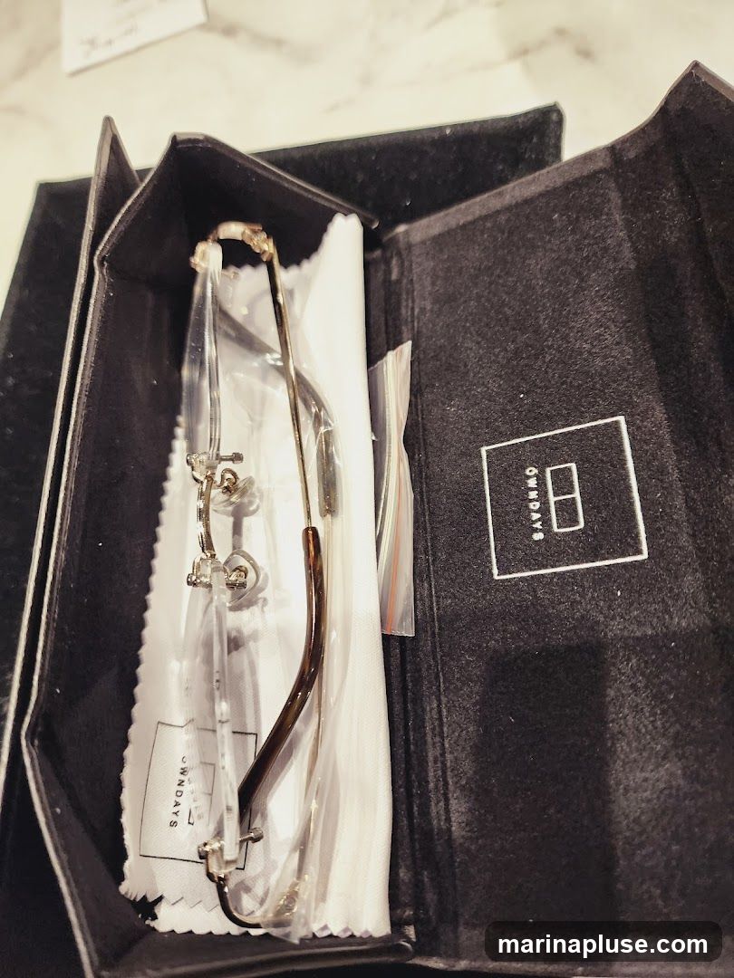 A pair of eyeglasses with clear lenses and brown temples are displayed inside a black, folded case. The eyeglasses are resting on a white cloth with a logo that reads "OWN DAYS". A small plastic bag containing what appears to be screws or nose pads is also visible. The inside of the case has a textured surface and a white square logo with the letters "HB" inside.