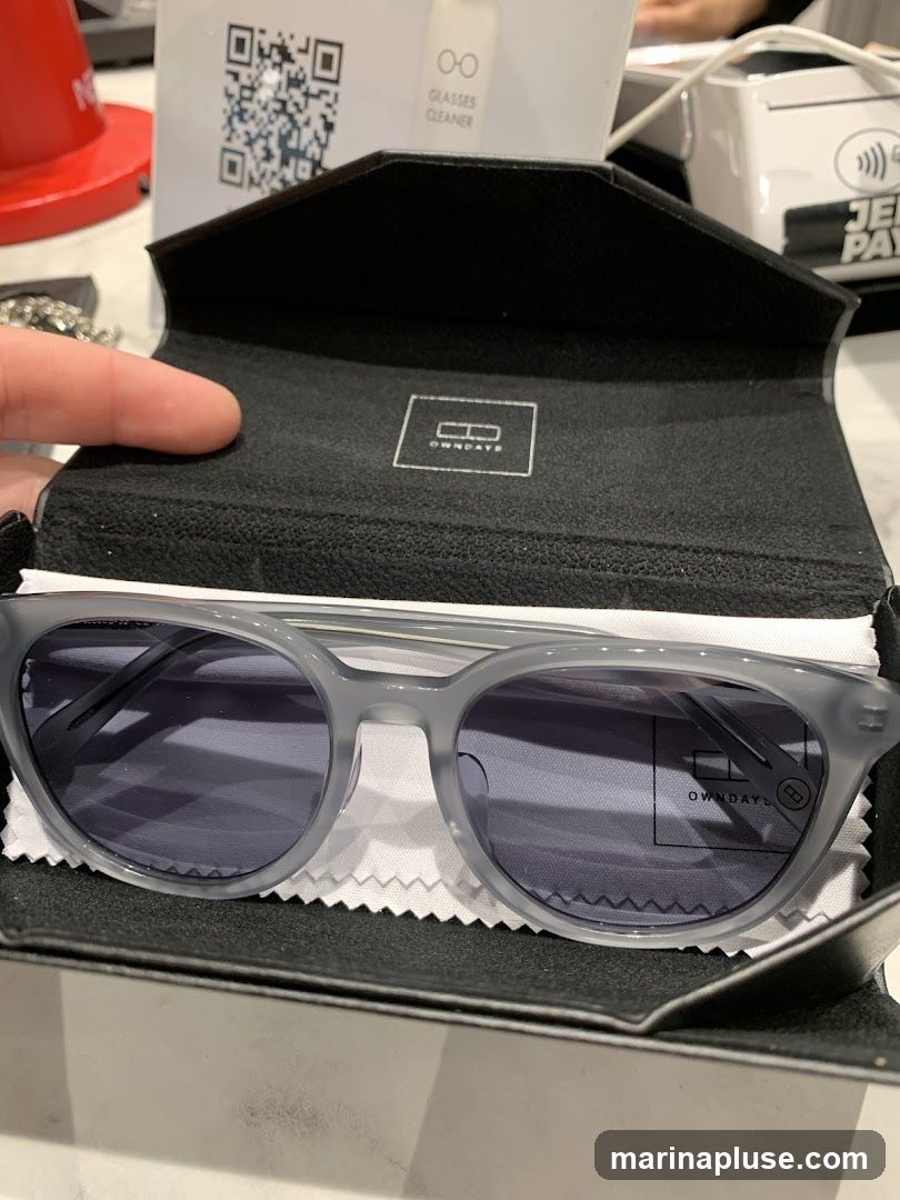 A hand holds open a black case revealing a pair of light gray sunglasses with purple lenses. The inside of the case has the "OWNDAYS" logo embossed. In the background, a QR code and a bottle labeled "GLASSES CLEANER" are visible.